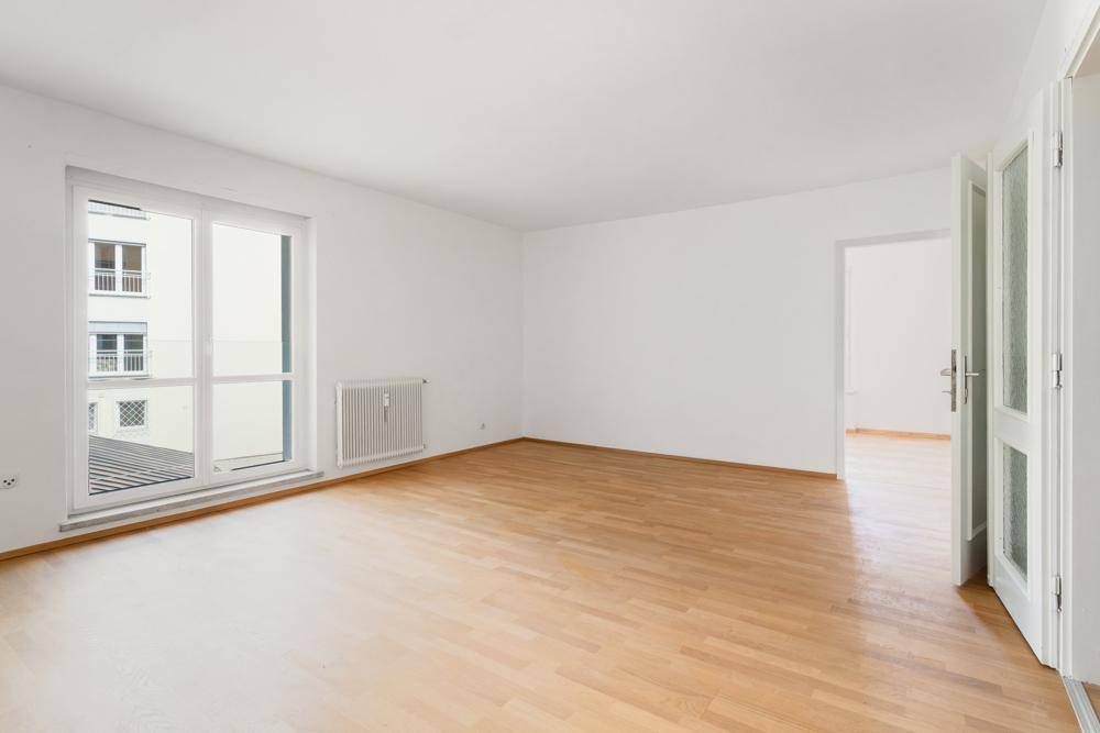 Heller, leerer Raum mit Parkettboden und großem Fenster, das viel Tageslicht hereinlässt.
