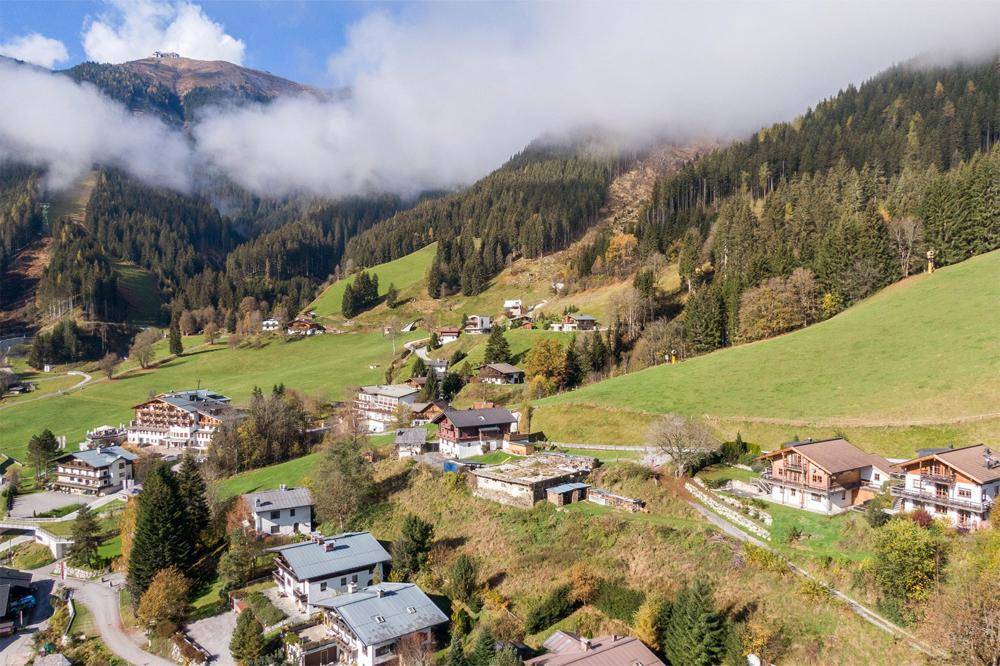Panoramablick auf ein Bergdorf in einem grünen Tal mit bewaldeten Hängen und Wolken.