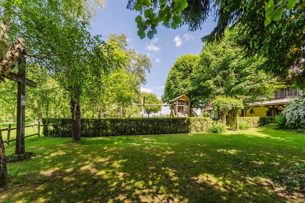 Idyllischer Gartenbereich mit sattgrünem Rasen, alten Bäumen und einem Holzkreuz, im Hintergrund ein Spielplatz.