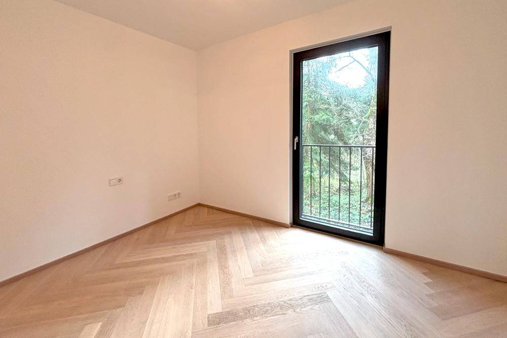 Leeres Zimmer mit hellem Fischgrätparkett und einem französischen Balkon mit Blick ins Grüne.