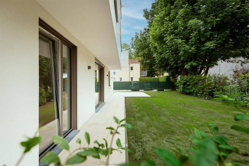 Gepflasterte Terrasse entlang des Hauses mit großen Fenstern und Zugang zum grünen Gartenbereich.