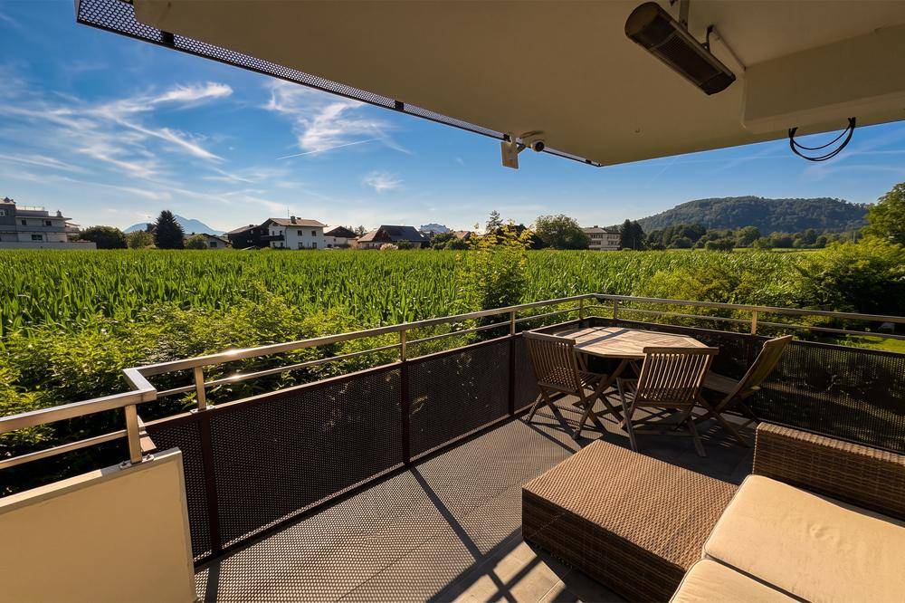 Sonniger Balkon mit bequemen Sitzgelegenheiten und weitem Blick über grüne Felder und die umliegende Berglandschaft.