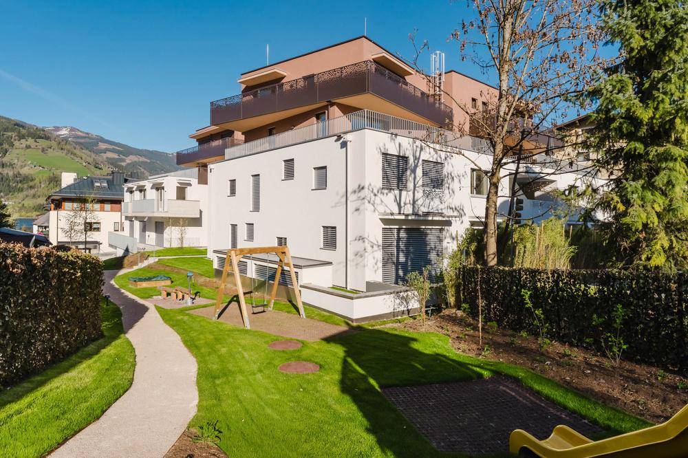 Modernes Wohngebäude mit gepflegtem Gemeinschaftsgarten, Kinderspielplatz und Balkonen vor Bergkulisse.