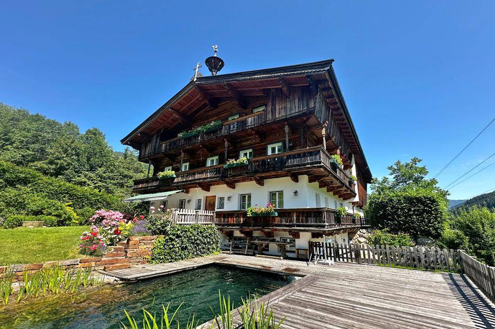 Traditionelles Holzhaus mit blühendem Garten, Naturpool und sonnigem Bergblick an einem klaren Sommertag.
