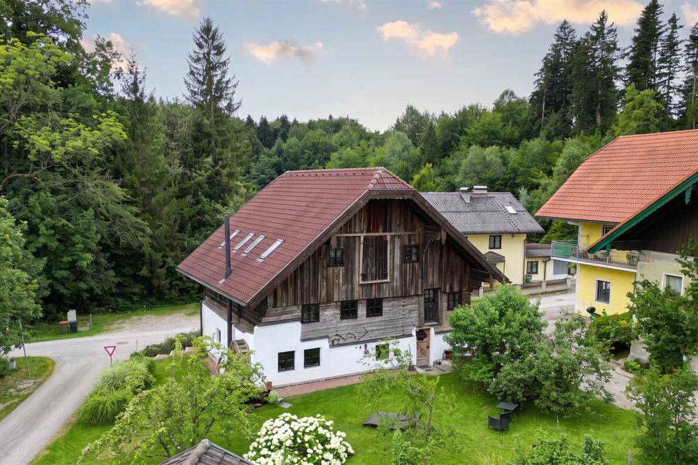 Traditionelles Holzhaus mit rotem Satteldach, umgeben von viel Grün und Bäumen in ländlicher Umgebung.