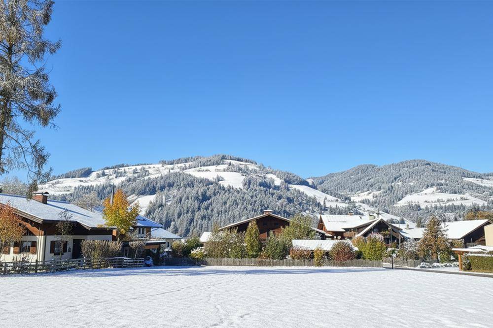 Ein idyllisches Dorf in einer verschneiten Berglandschaft, umgeben von schneebedeckten Bäumen und Hügeln unter klarem Himmel.