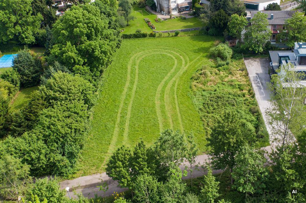 Luftaufnahme eines großen grünen Grundstücks mit gemähten Mustern, umgeben von Bäumen und Wohngebäuden, inklusive Tennisplatz.