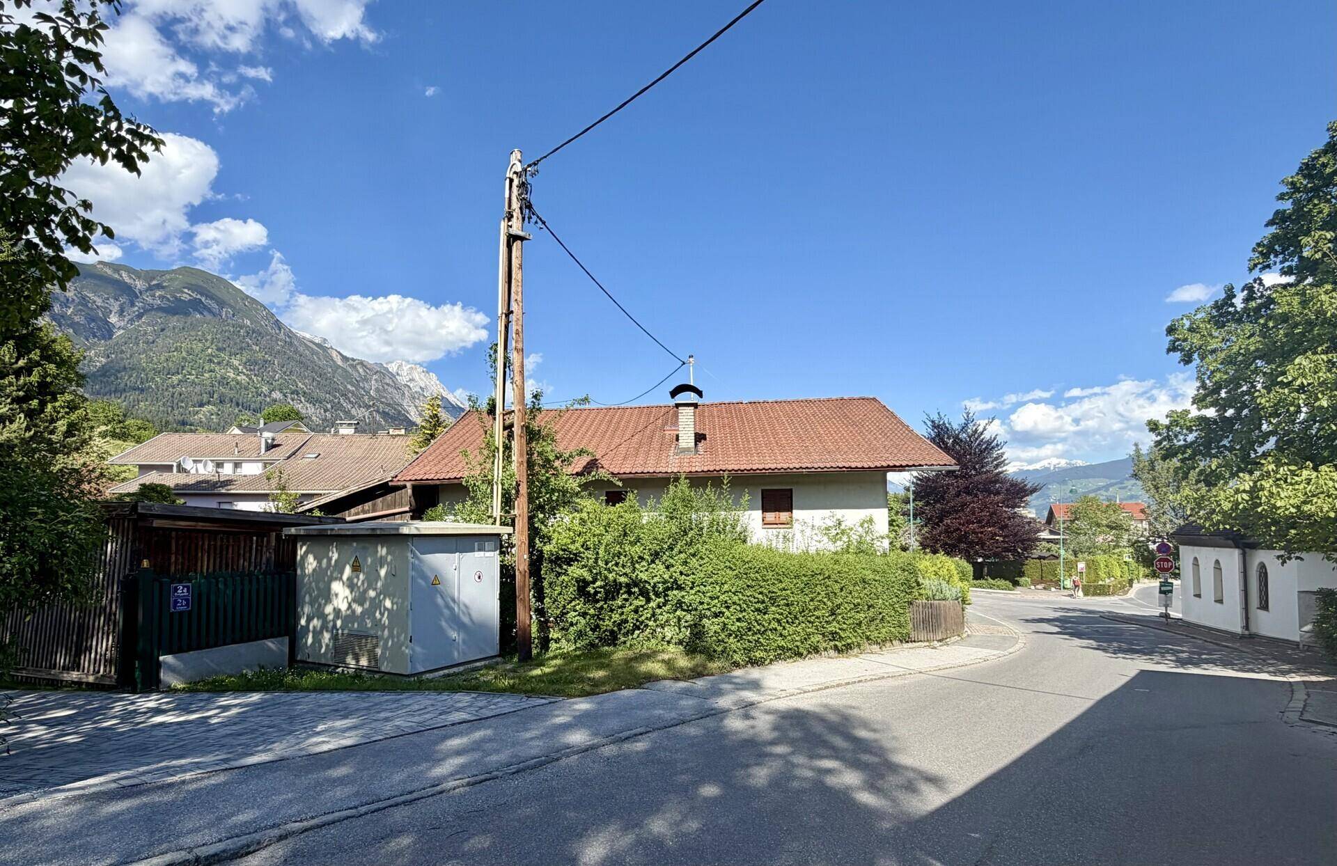 Ruhige Wohnstraße mit einem Haus im Hintergrund, umgeben von Bäumen und Bergen.