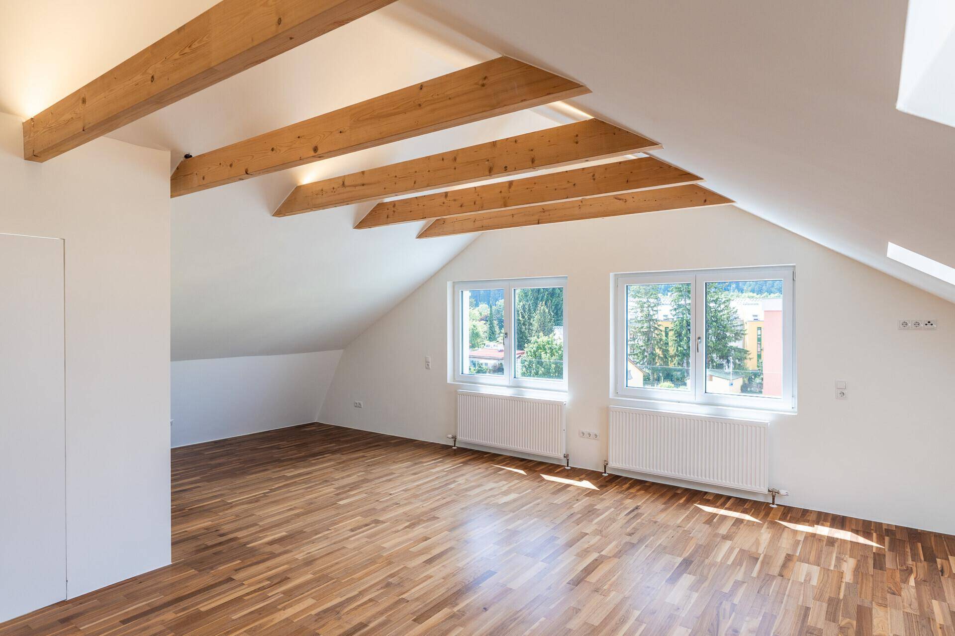 Geräumiges Dachgeschosszimmer mit Holzbalken, Parkettboden und großen Fenstern, die einen Blick ins Grüne ermöglichen.
