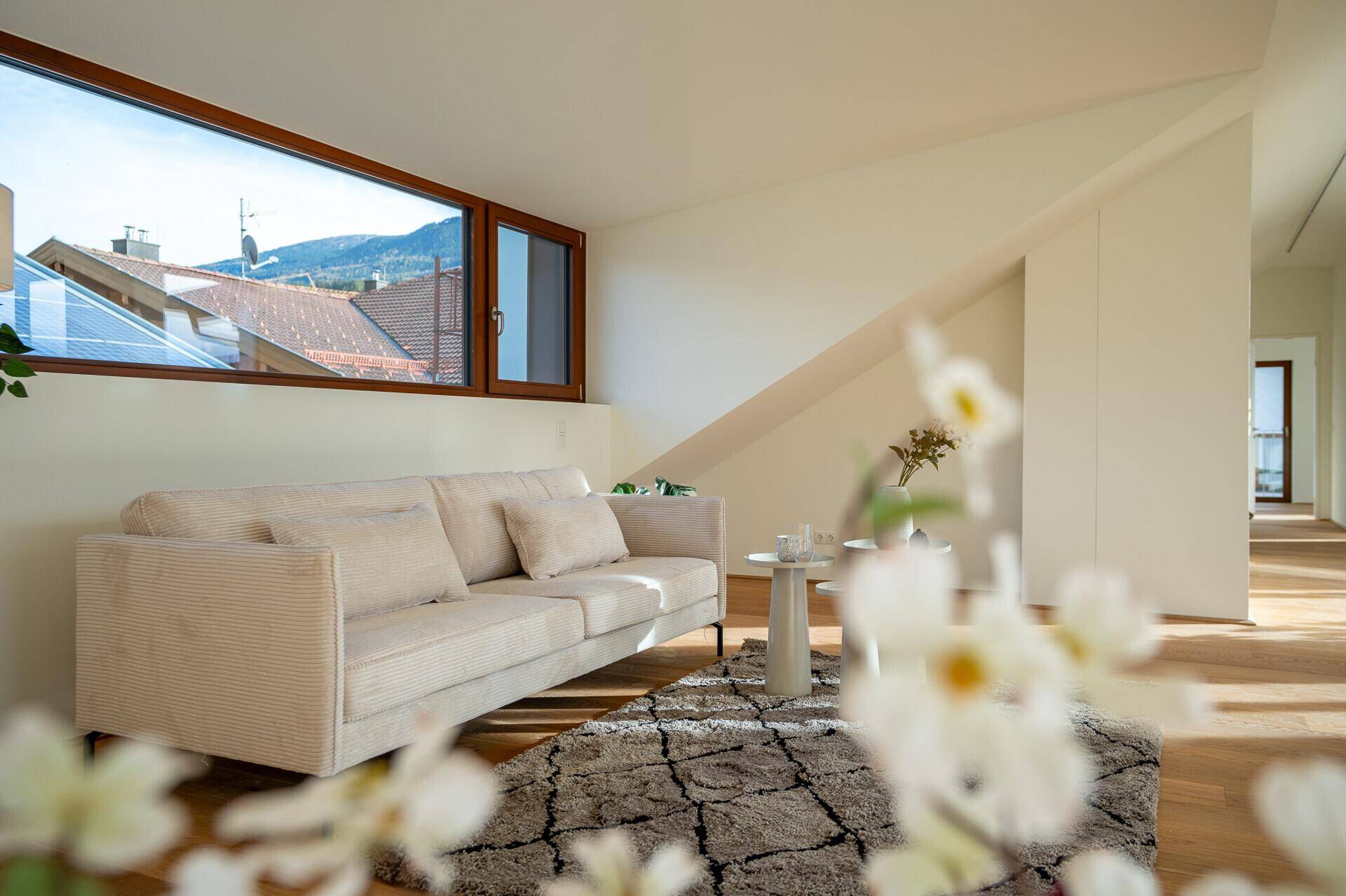 Gemütliches Wohnzimmer mit beigem Sofa, Holzboden und einem breiten Fenster unter der Dachschräge.