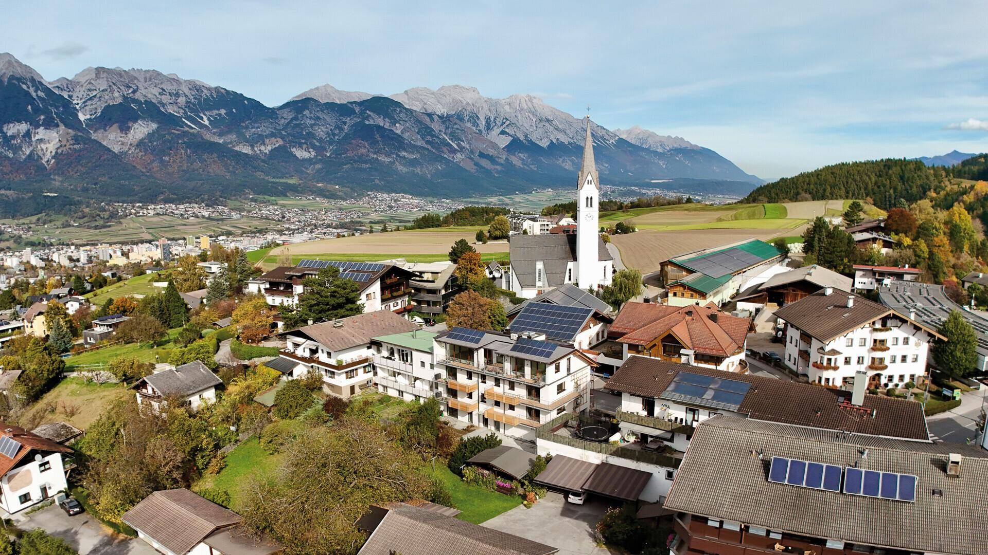 Malerische Luftaufnahme eines Dorfes mit Kirche, umgeben von grünen Feldern und majestätischen Bergen.