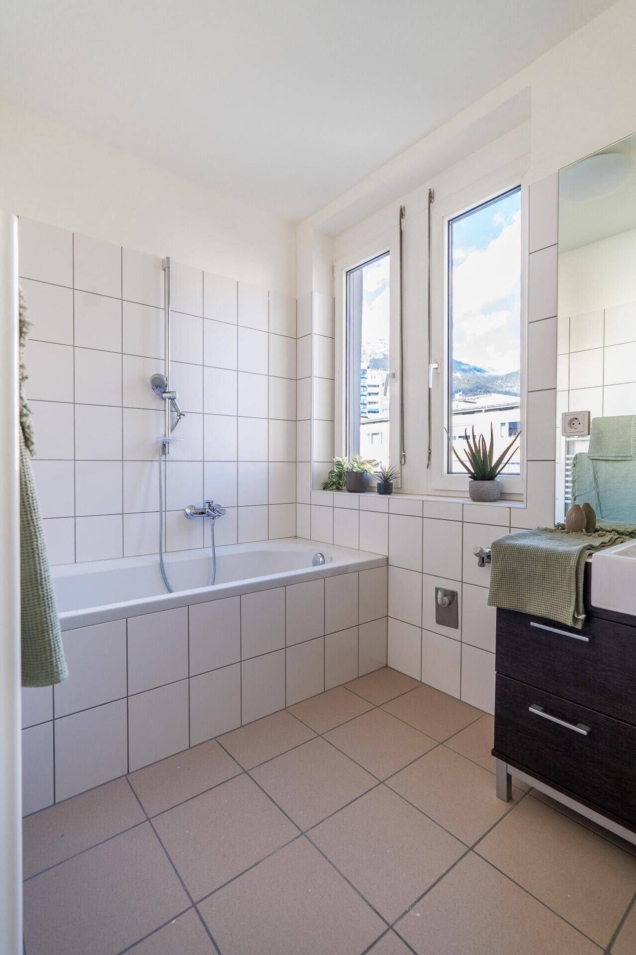 Helles Badezimmer mit Badewanne, weißen Fliesen und Fenster mit Bergblick.