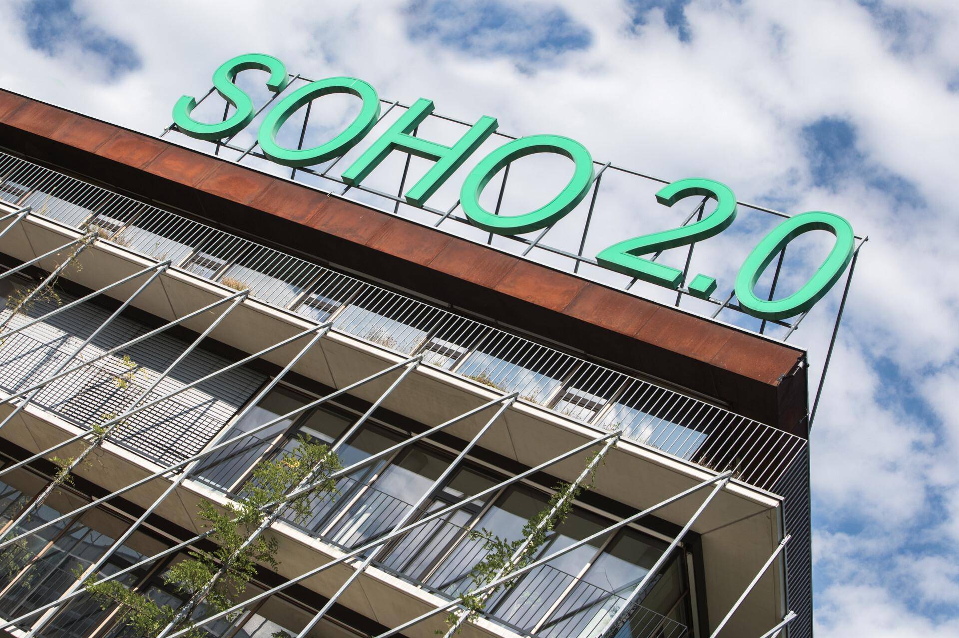 Nahaufnahme der modernen Gebäudefassade mit dem markanten "SOHO 2.0" Schriftzug vor bewölktem Himmel.