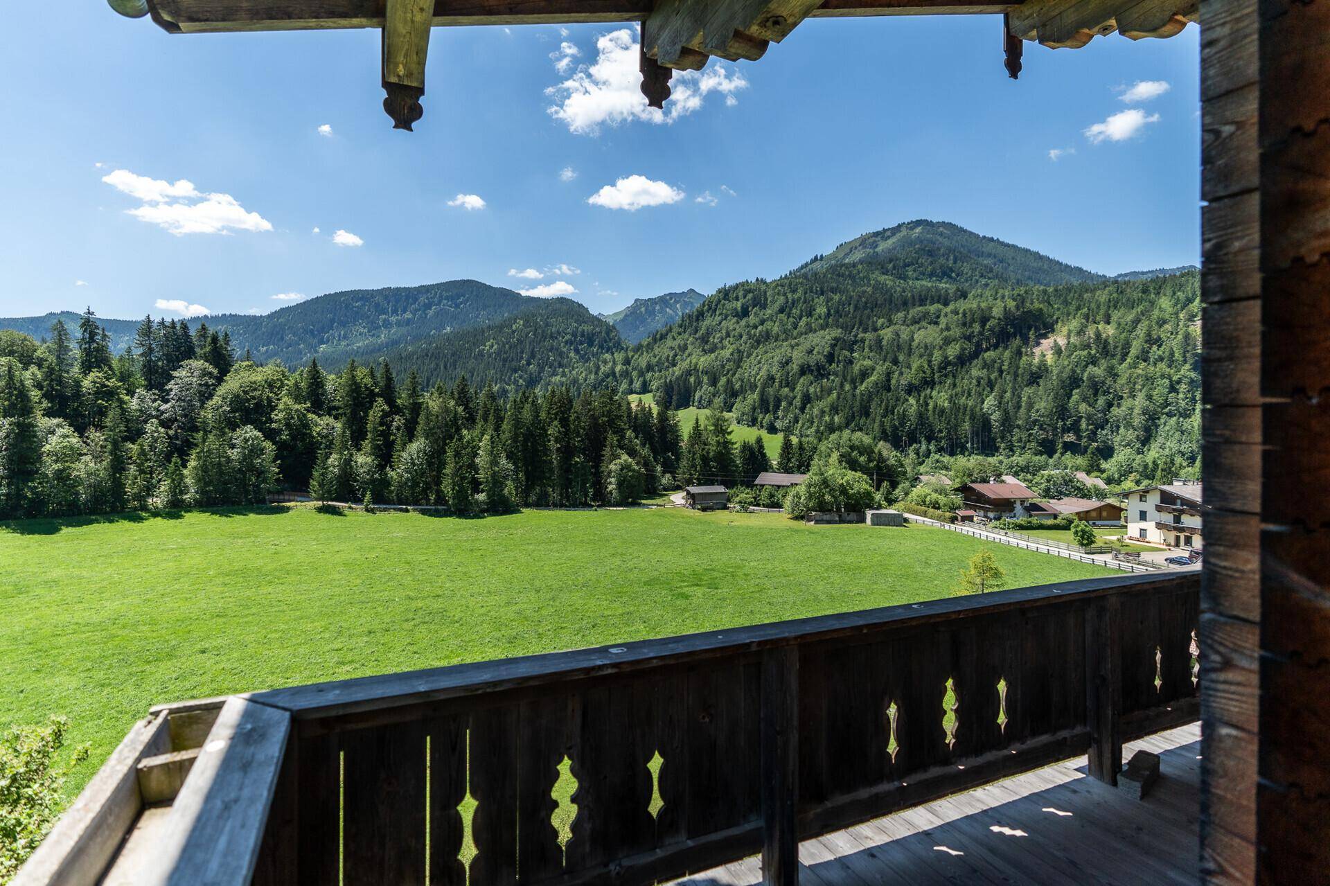 Panoramablick vom Holzbalkon über weite grüne Felder und majestätische Berge unter blauem Himmel.