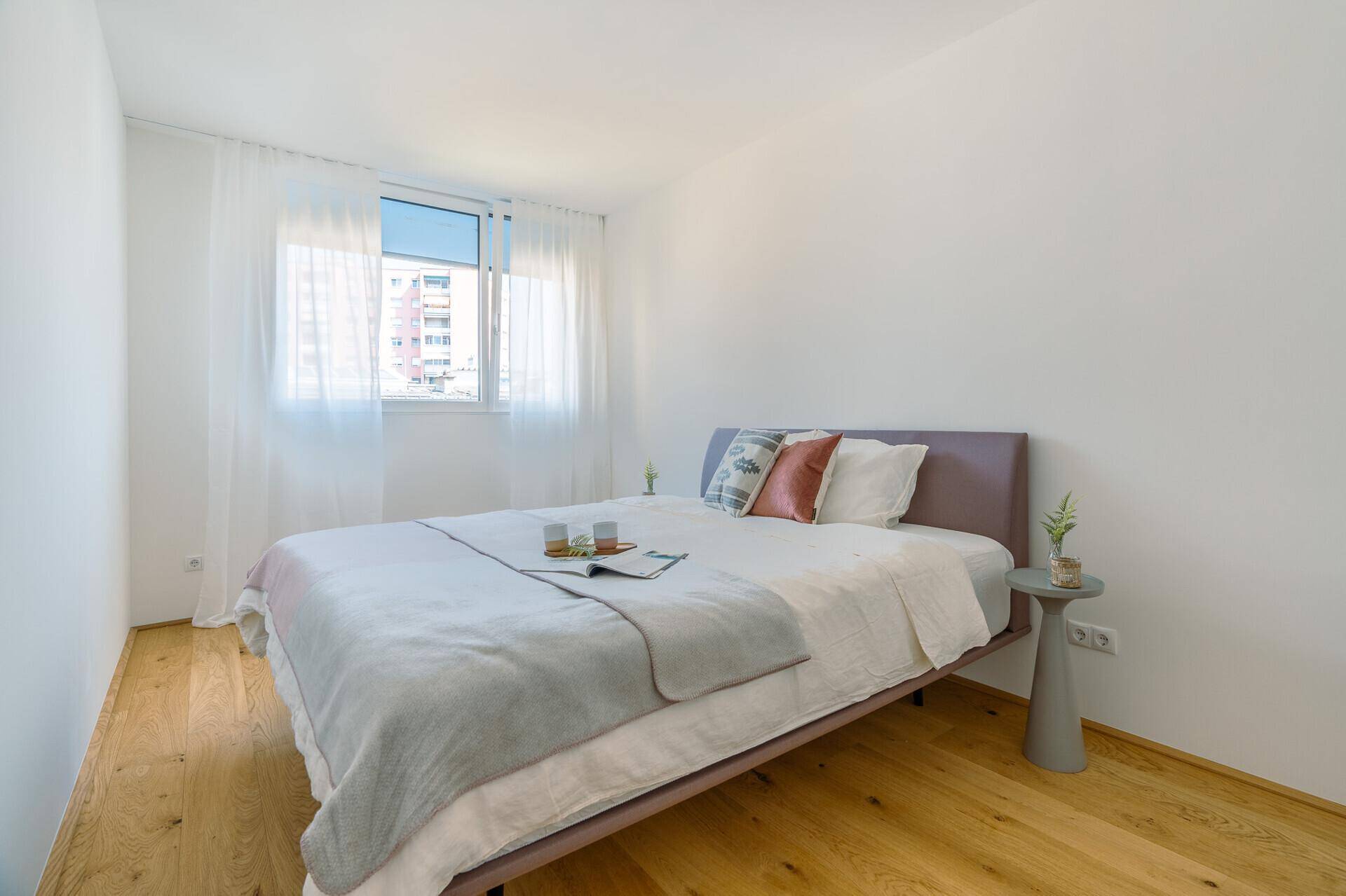 Helles Schlafzimmer mit Holzboden, weiß gestrichenen Wänden und einem Fenster mit Vorhängen.
