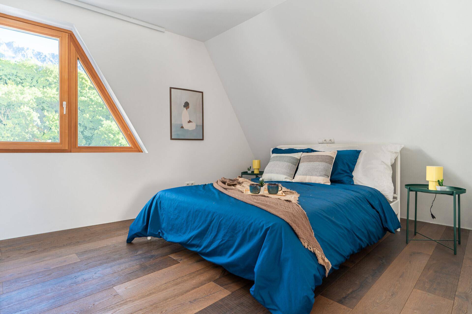Schlafzimmer mit blauem Bett, Holzboden und dreieckigem Fenster mit Blick ins Grüne.