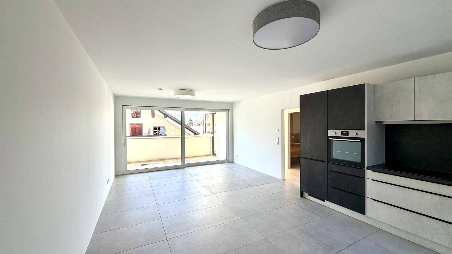 Heller offener Wohnbereich mit moderner Küchenzeile, Fliesenboden und Zugang zum Balkon.