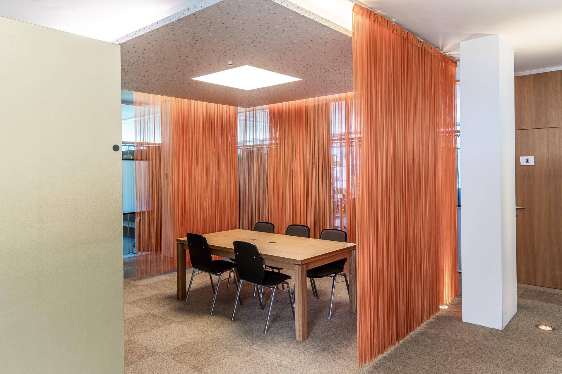 Moderner Besprechungsraum mit Holztisch und orangefarbenen Fadenvorhängen als Raumteiler.