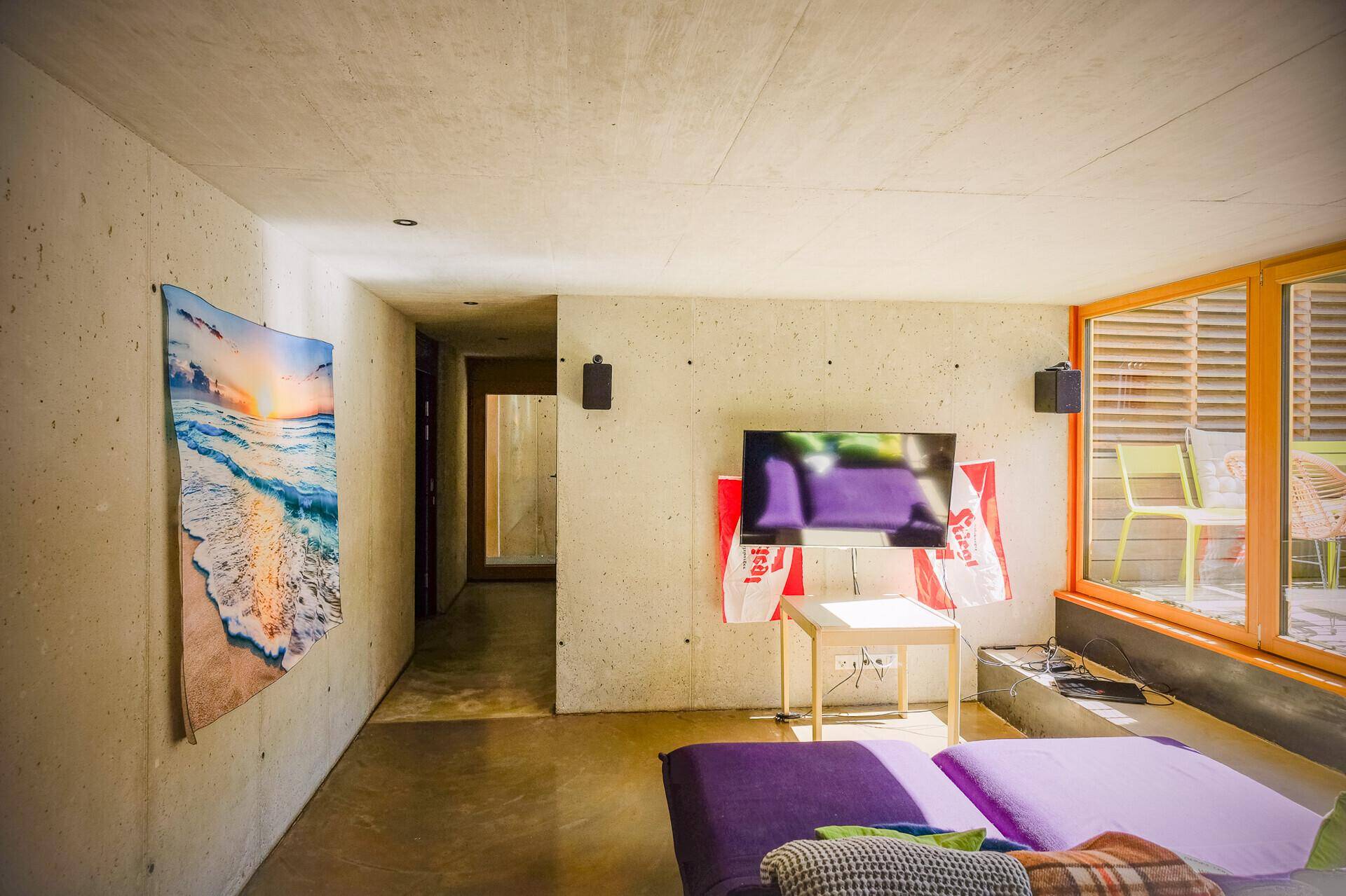 Moderner Wohnbereich mit Sichtbetonwänden, großem Fenster und gemütlicher Sitzgelegenheit.