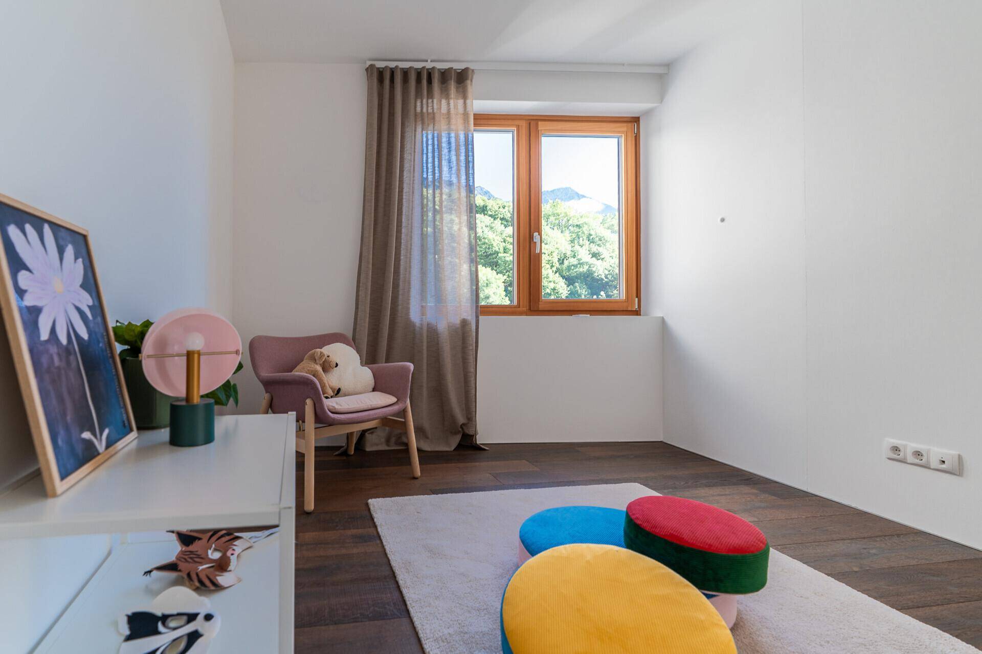 Helles Zimmer mit Holzboden, bunten Sitzkissen und Fenster mit Bergblick.
