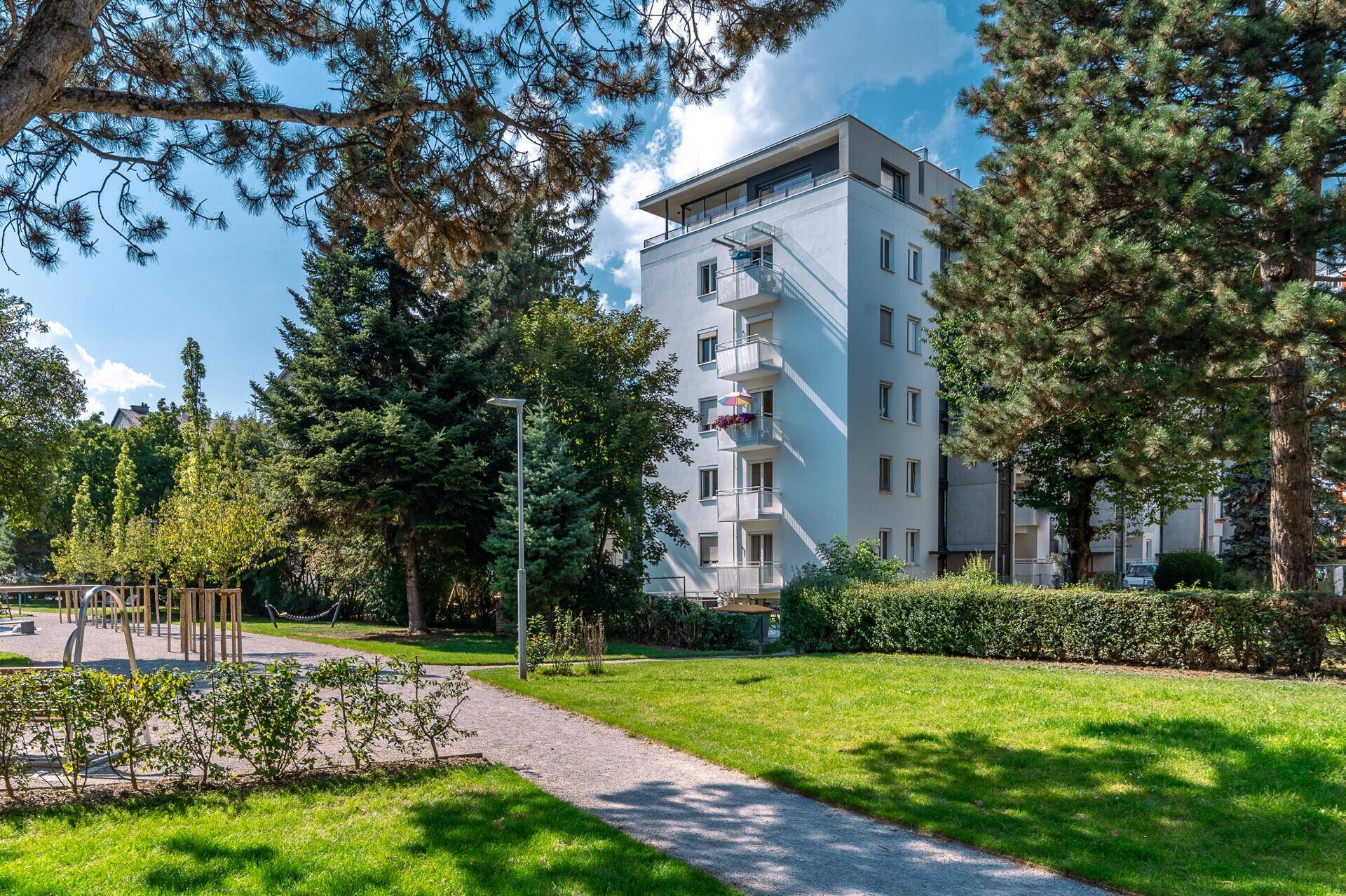 Modernes Apartmentgebäude in einer grünen Parkanlage mit Bäumen und Spazierwegen.