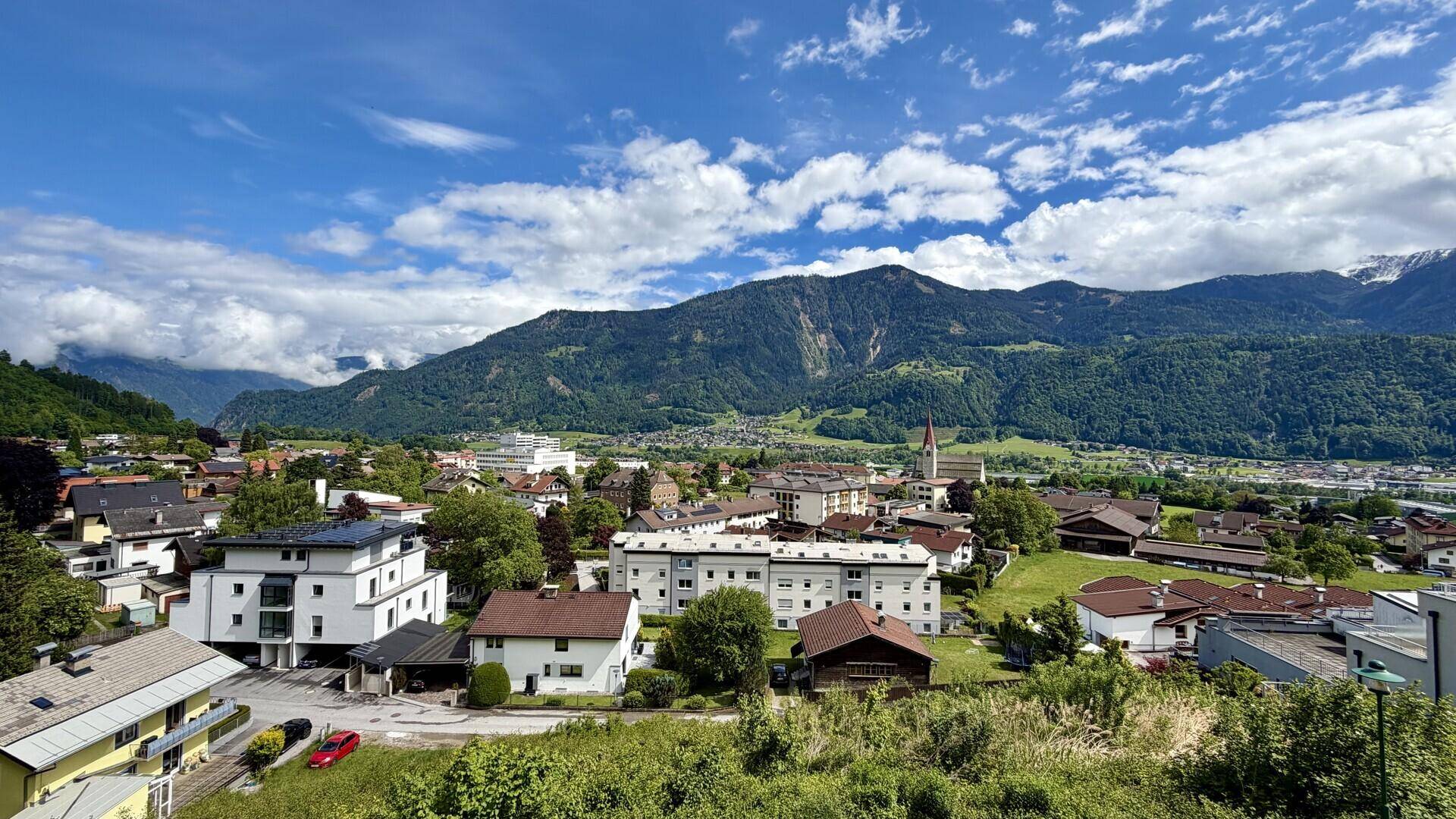 Weitläufiger Blick über eine malerische Stadtlandschaft mit grünen Hügeln und majestätischen Bergen im Hintergrund.