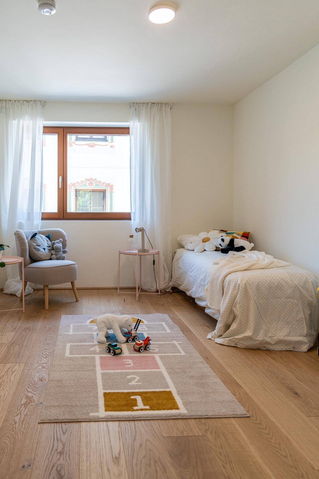 Freundliches Kinderzimmer mit Holzboden, Einzelbett und einem bunten Spielteppich, gut beleuchtet.