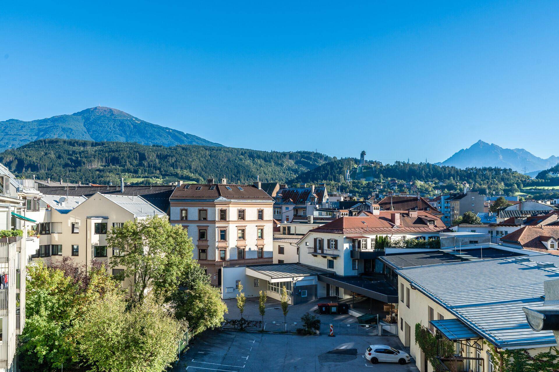 Weitläufiger Blick über eine Stadtlandschaft mit Gebäuden und grünen Bergen am Horizont.