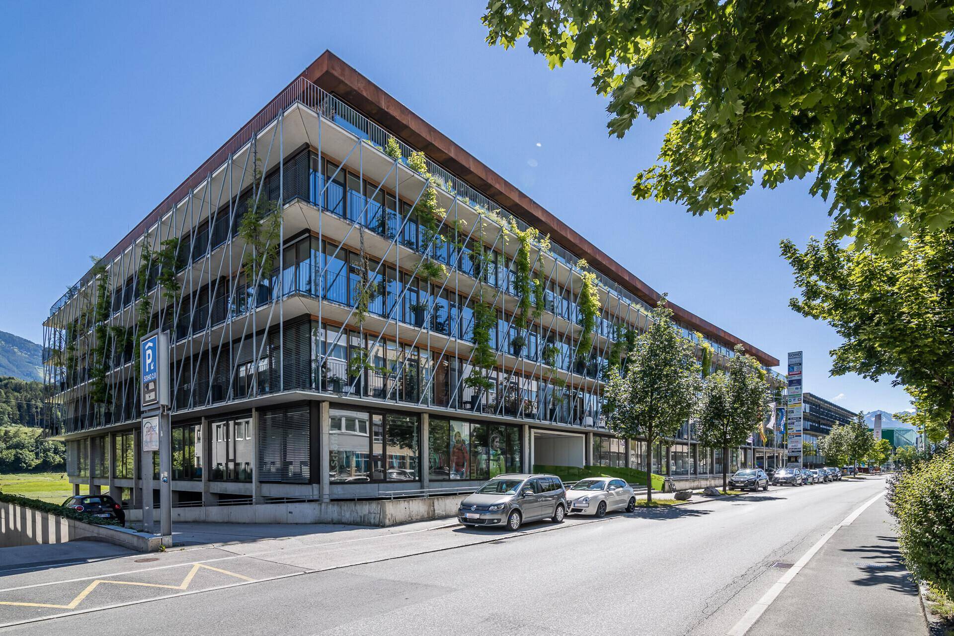 Modernes Bürogebäude an einer Straße mit geparkten Autos und viel Grün.