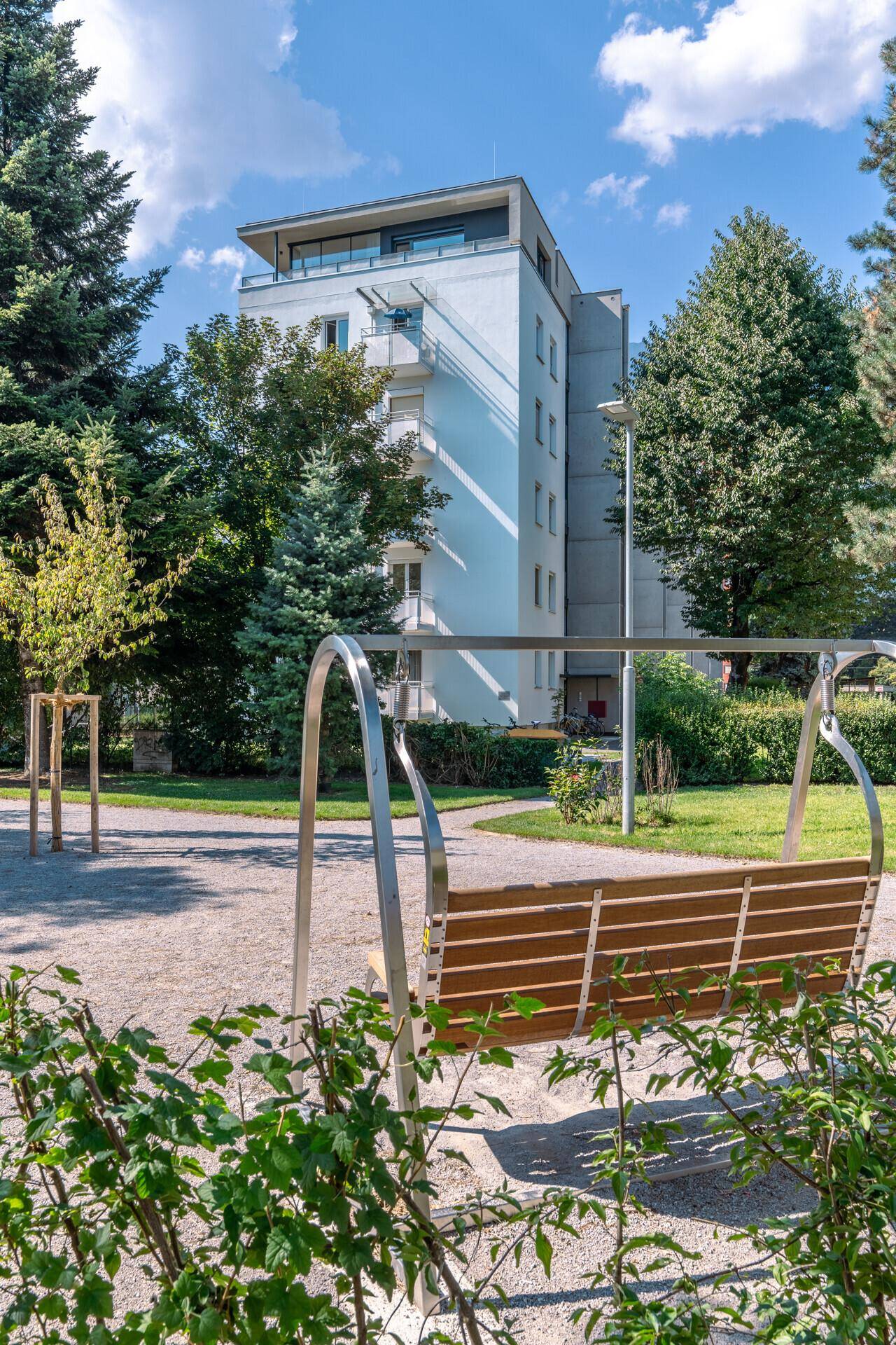 Parkanlage mit moderner Schaukelbank und Blick auf das Apartmentgebäude im Hintergrund.