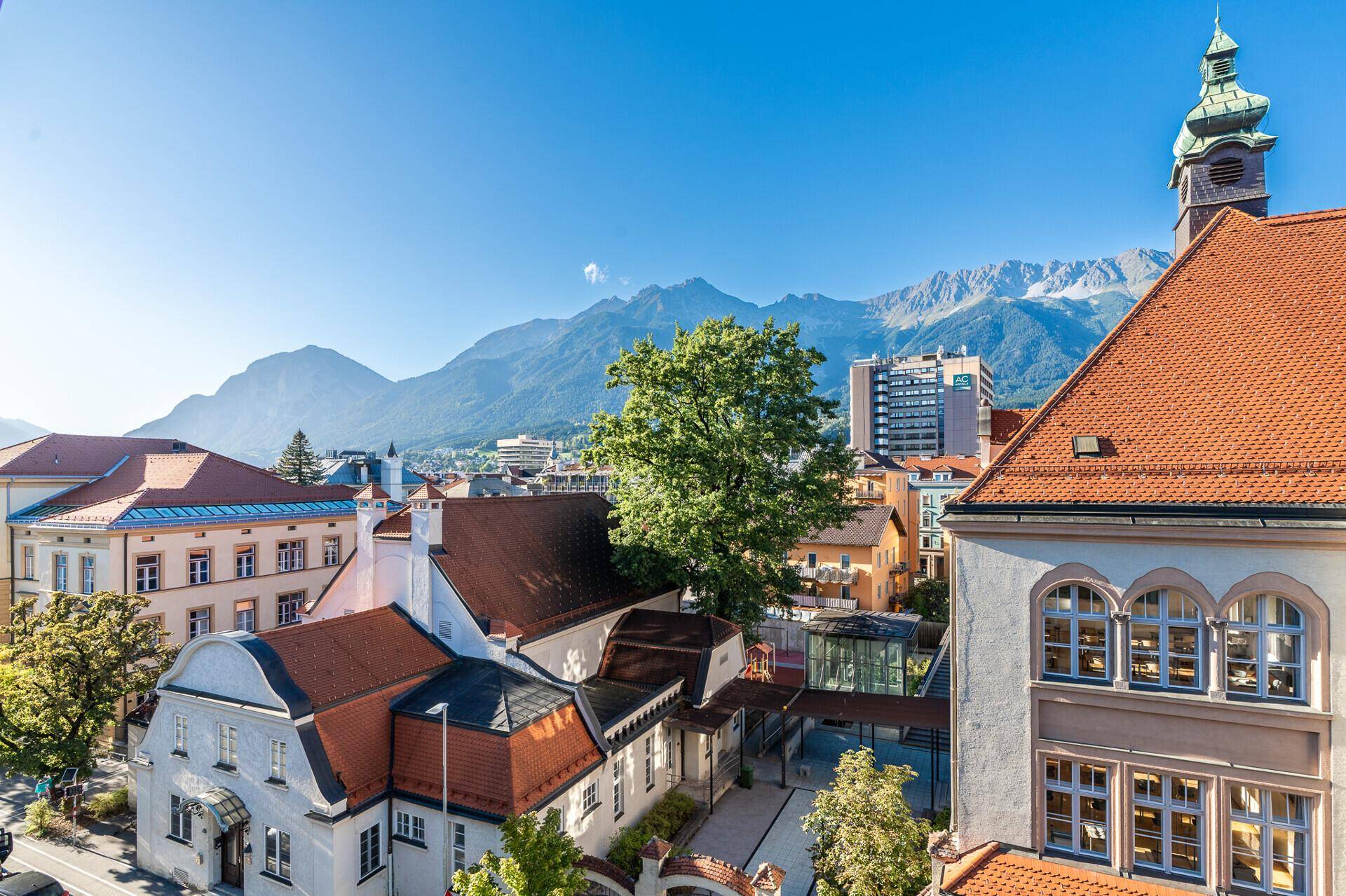 Panoramablick über die Stadt mit roten Dächern und majestätischen Bergen im Hintergrund unter blauem Himmel.