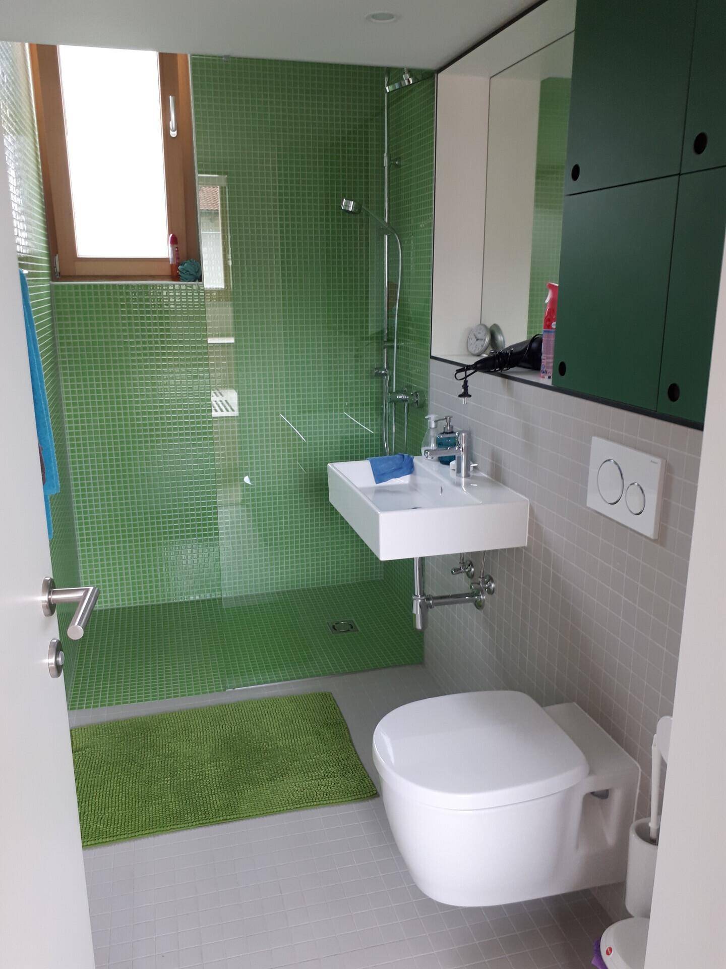Helles Badezimmer mit grünen Mosaikfliesen im Duschbereich, wandhängendem WC und Fenster.