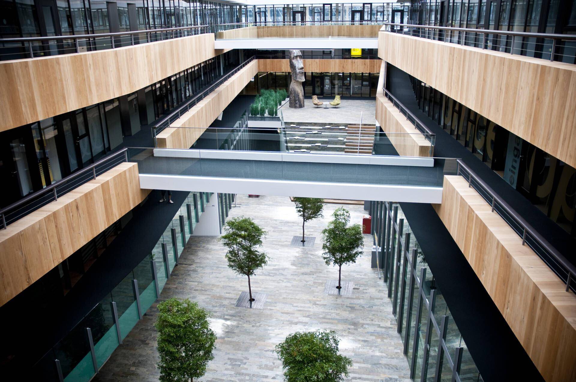 Großzügiges, mehrstöckiges Atrium mit Bäumen, Holzverkleidungen und gläsernen Übergängen.