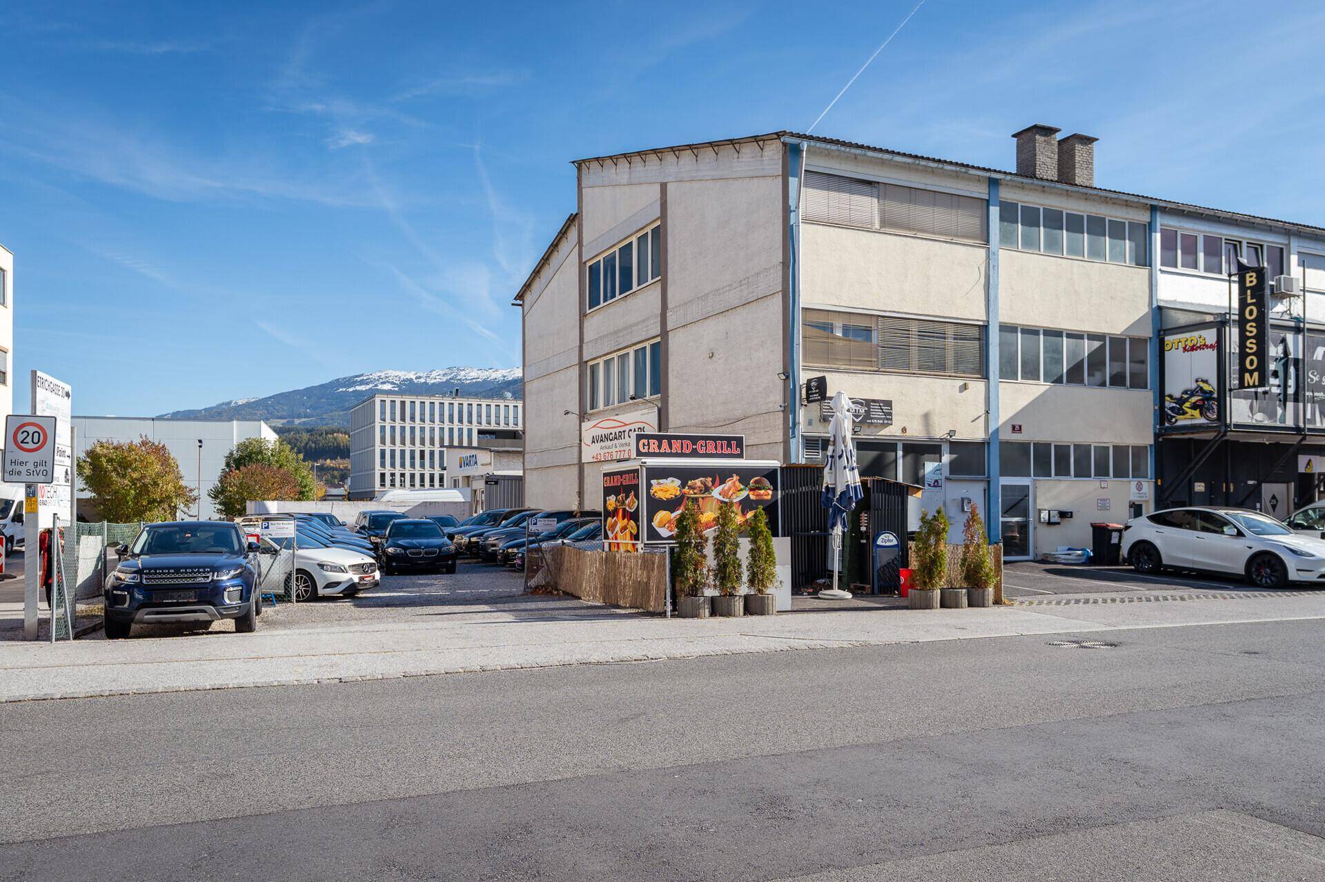Gebäude mit Geschäftsfassaden und Parkplätzen an einer Straße in einem städtischen Umfeld.