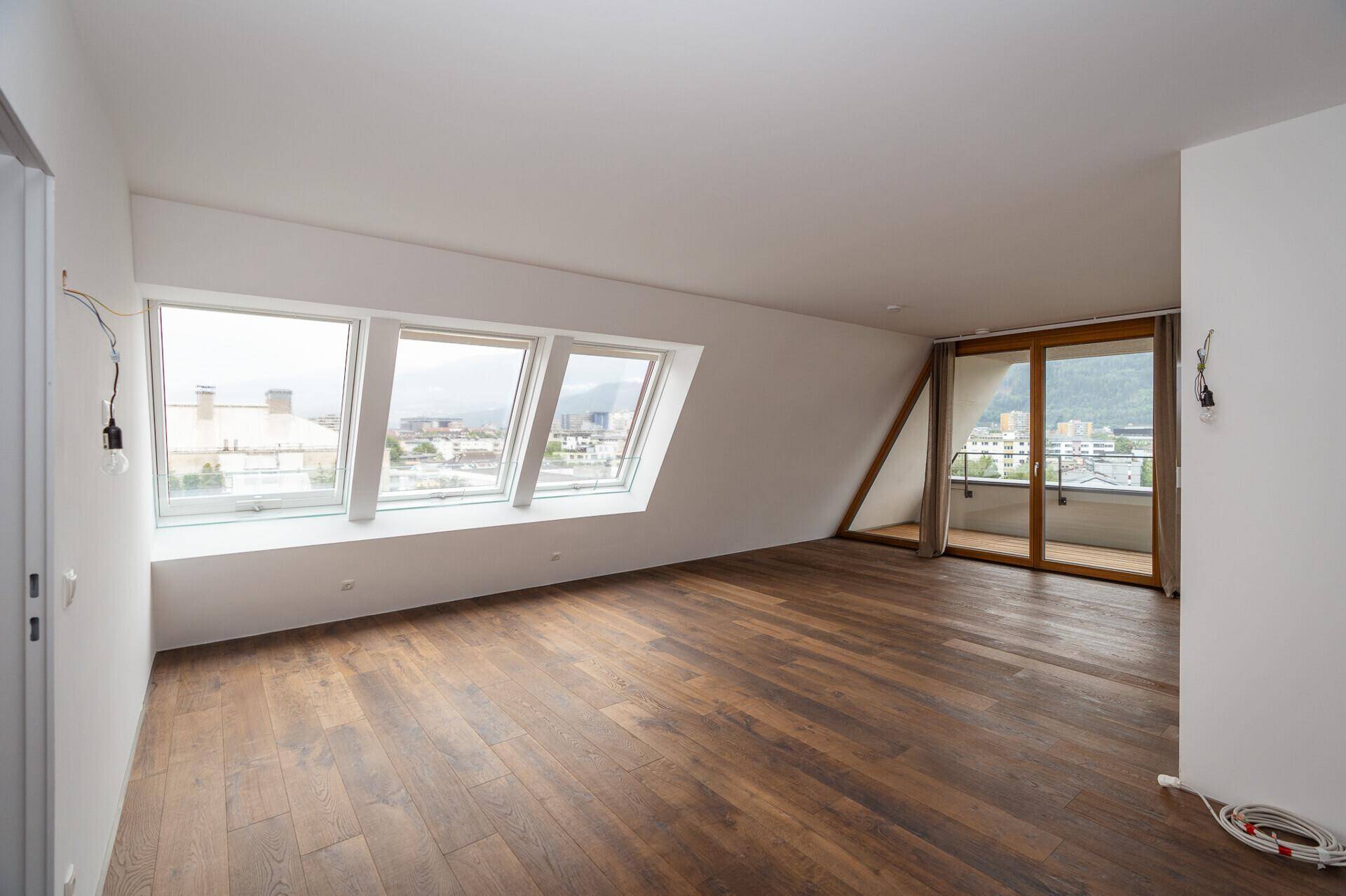 Heller Raum mit Holzboden, Dachschräge, großen Fenstern und Zugang zu einem Balkon mit Stadt- und Bergblick.