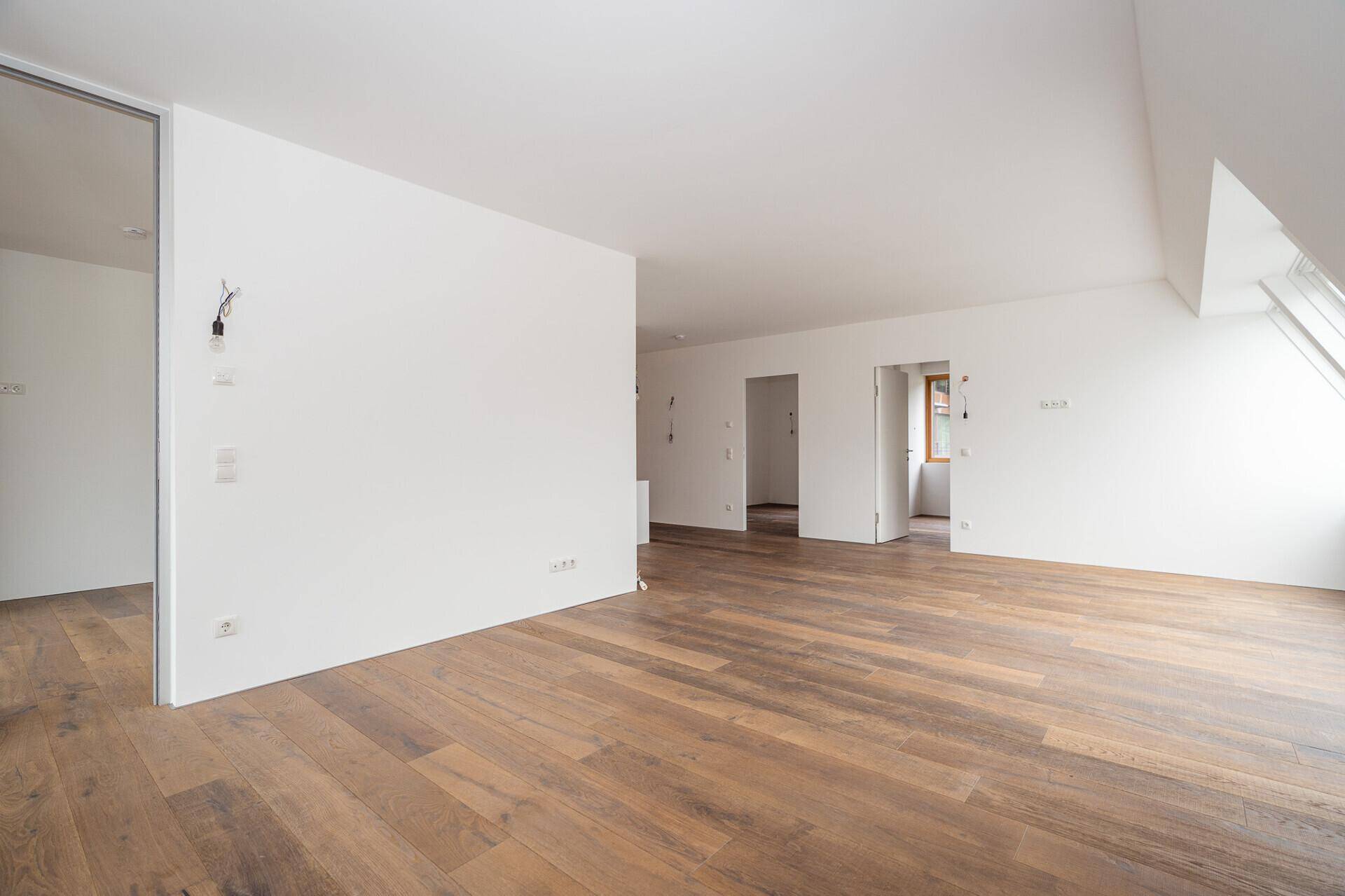 Heller, leerer Raum mit Holzboden, weißen Wänden und mehreren Türöffnungen.