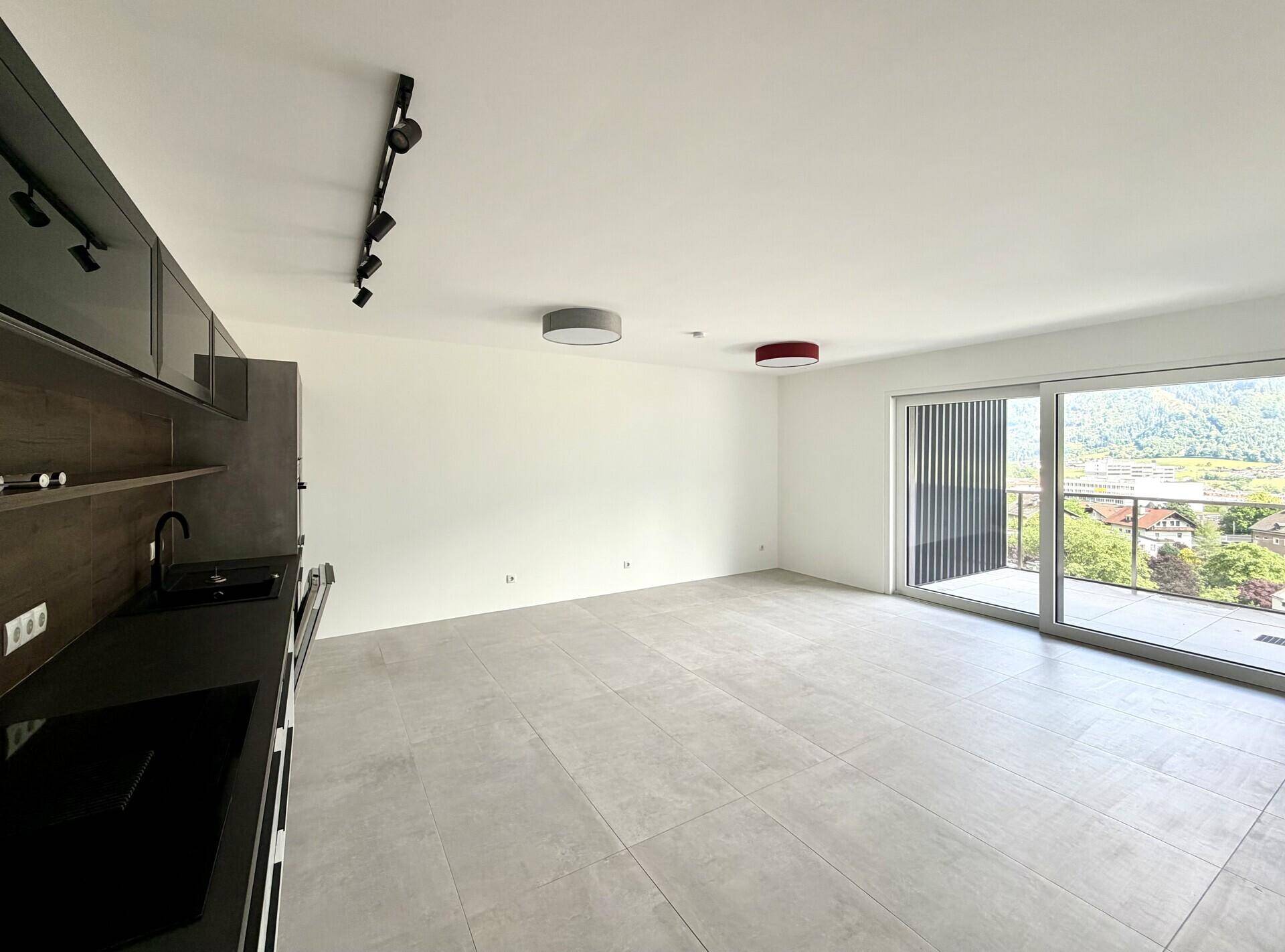 Heller offener Wohnbereich mit integrierter Küche, Fliesenboden und Zugang zum Balkon mit Bergblick.