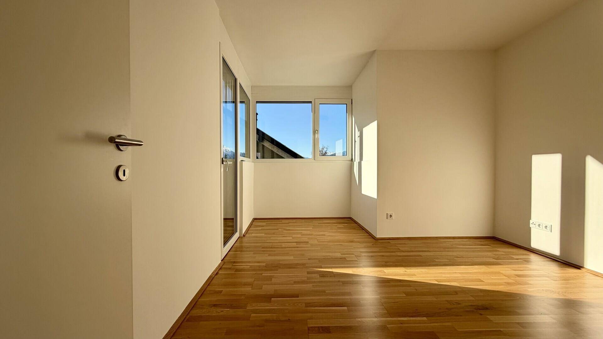 Lichtdurchflutetes Zimmer mit Holzboden und großen Fenstern, die viel Sonnenlicht hereinlassen.