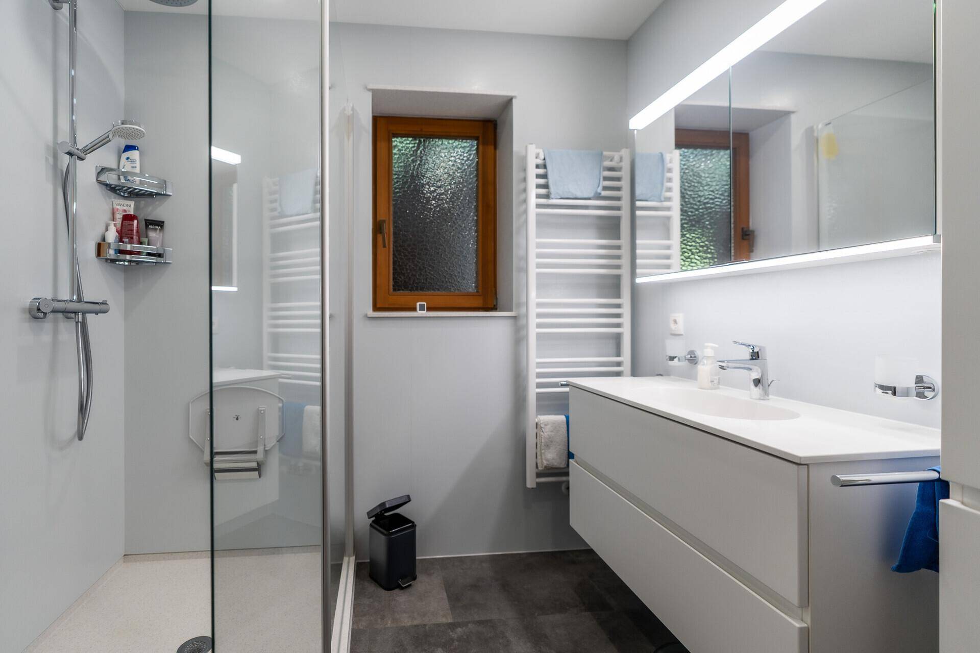 Modernes Badezimmer mit ebenerdiger Dusche, Einzelwaschbecken, beheiztem Handtuchhalter und Fenster.