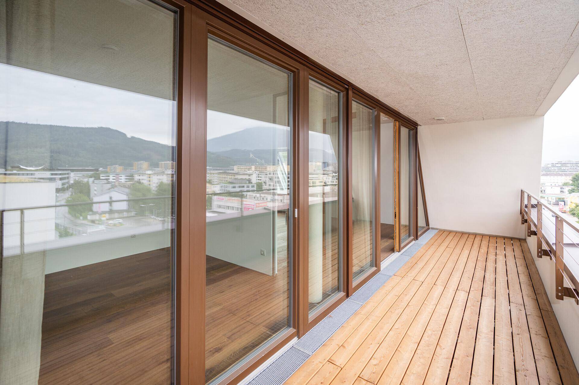 Geräumiger Balkon mit Holzbelag und Glasgeländer, bietet einen weiten Blick auf die Berge und die Stadt.