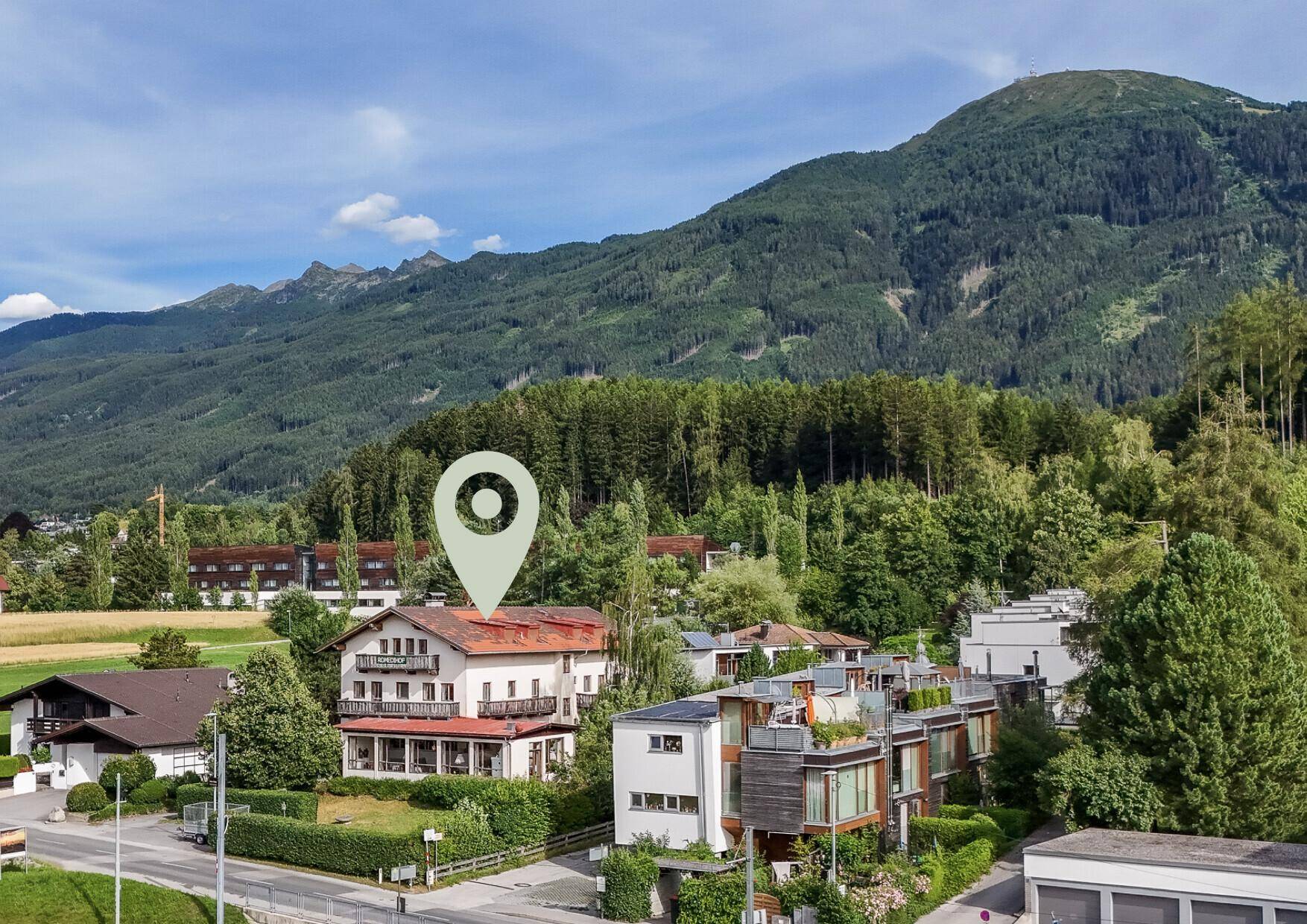 Luftaufnahme eines Gebäudes in grüner Umgebung mit bewaldeten Bergen im Hintergrund.
