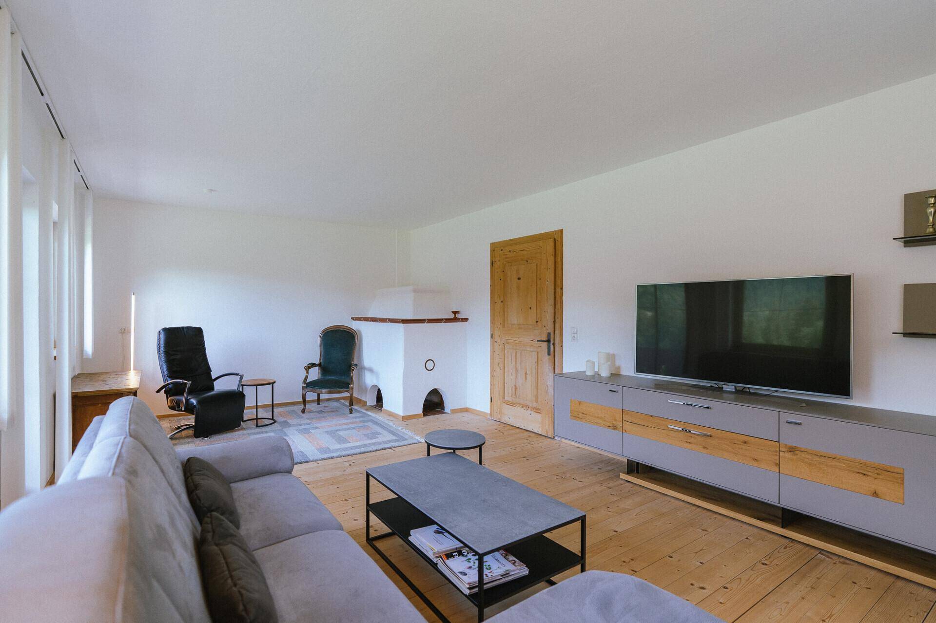 Helles Wohnzimmer mit Holzboden, modernem Sofa, TV-Möbel und einem traditionellen Kachelofen.