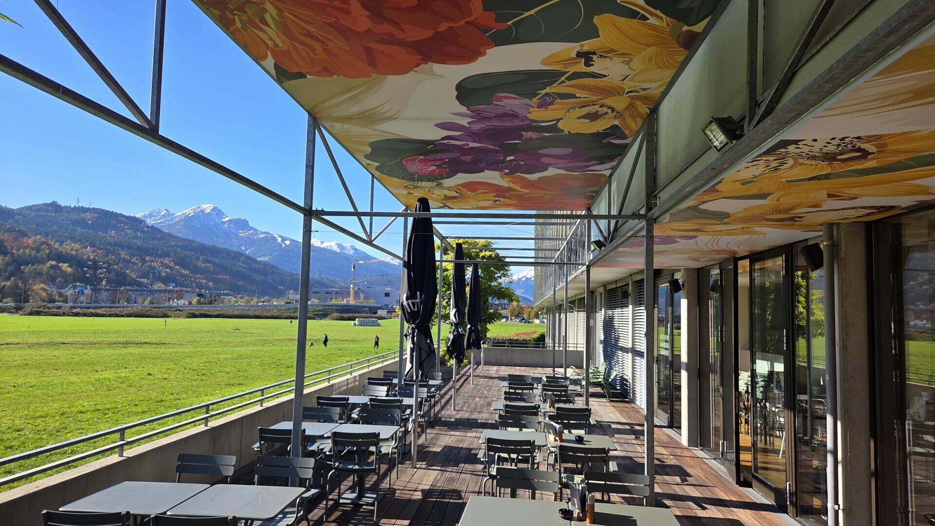 Große Außenterrasse eines Cafés mit Holzboden, vielen Tischen und einem beeindruckenden Bergblick.