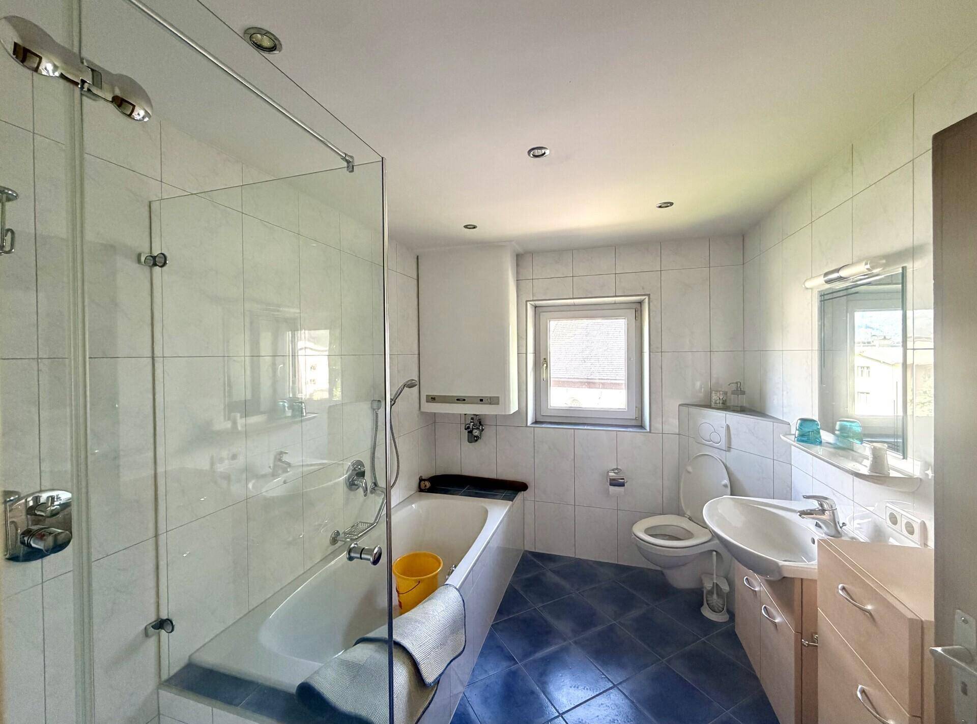 Helles Badezimmer mit weißen Fliesen, blauen Bodenfliesen, Badewanne und Duschabtrennung.