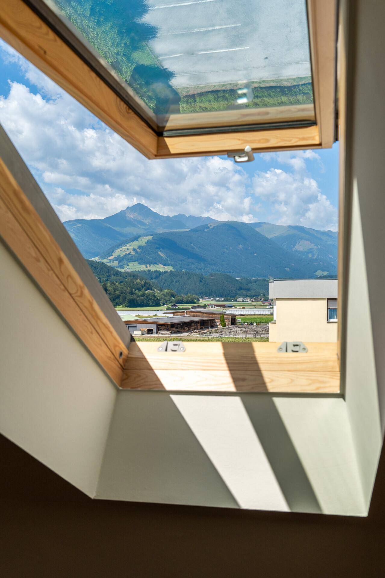 Atemberaubender Bergblick durch ein geöffnetes Dachfenster, umgeben von grüner Landschaft.