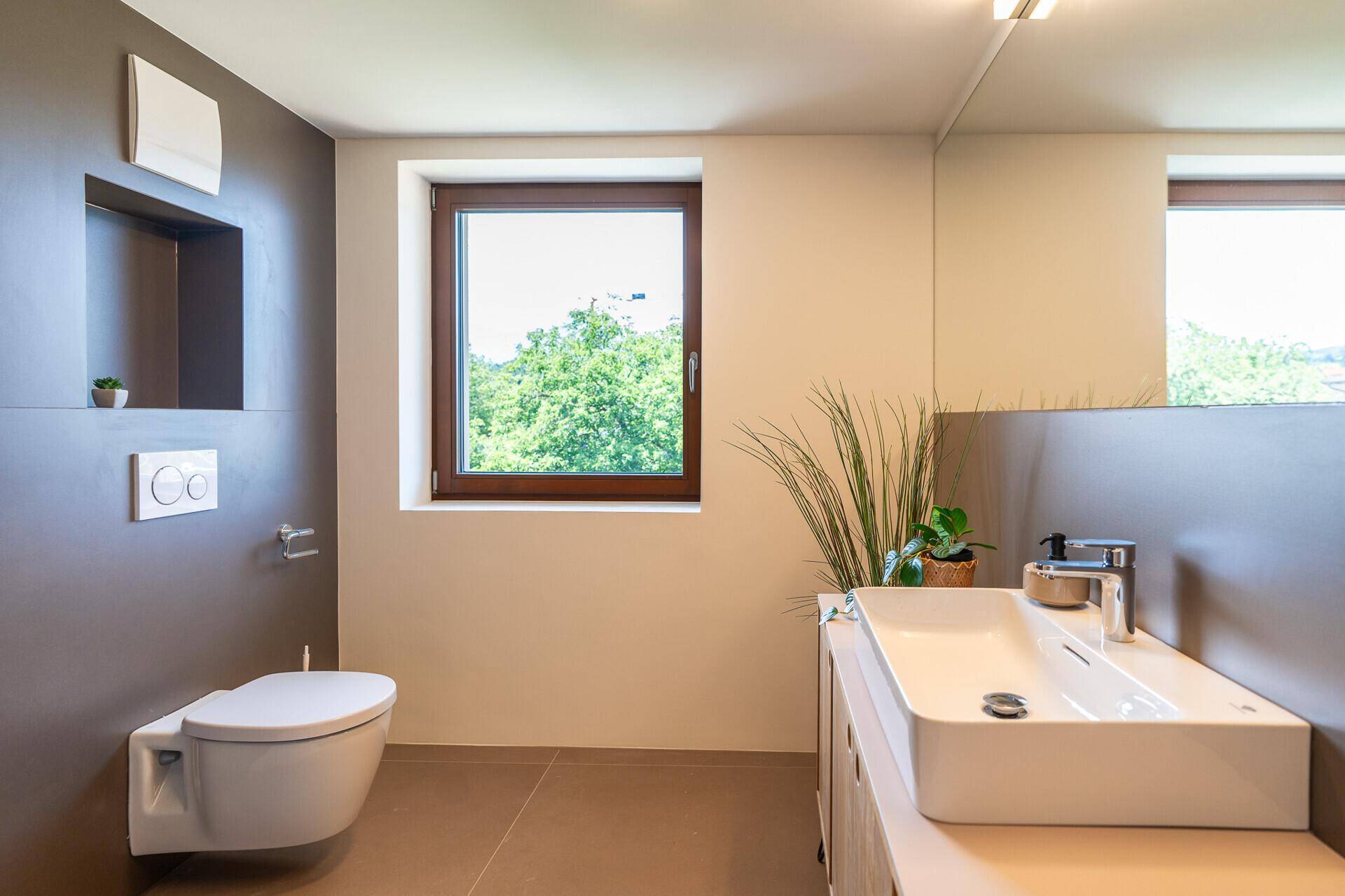 Modernes Badezimmer mit Wand-WC, Aufsatzwaschbecken und Fenster, in neutralen Grau- und Weißtönen gehalten.