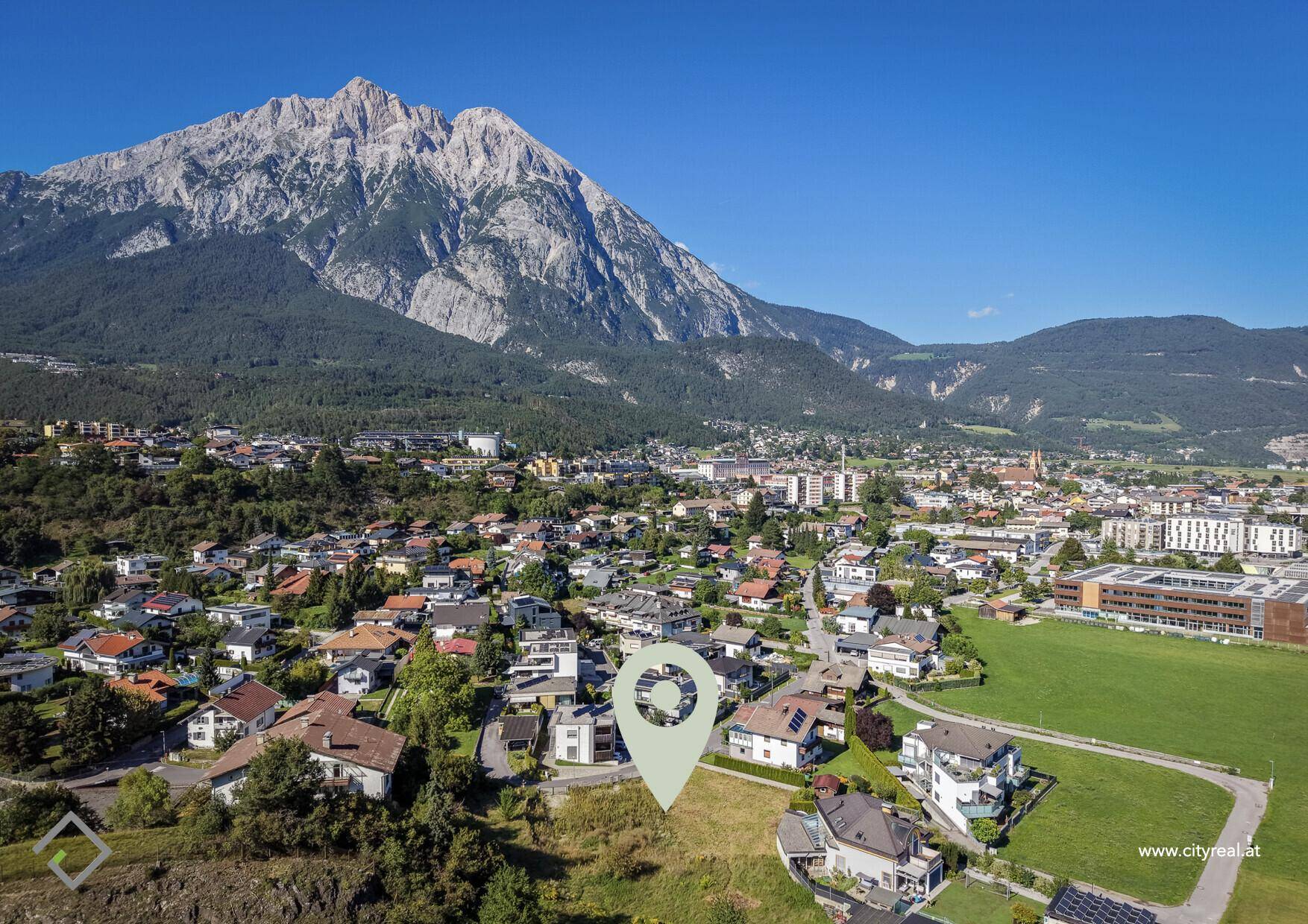 Panoramablick aus der Vogelperspektive auf ein Wohngebiet mit markiertem Grundstück und majestätischer Bergkulisse.