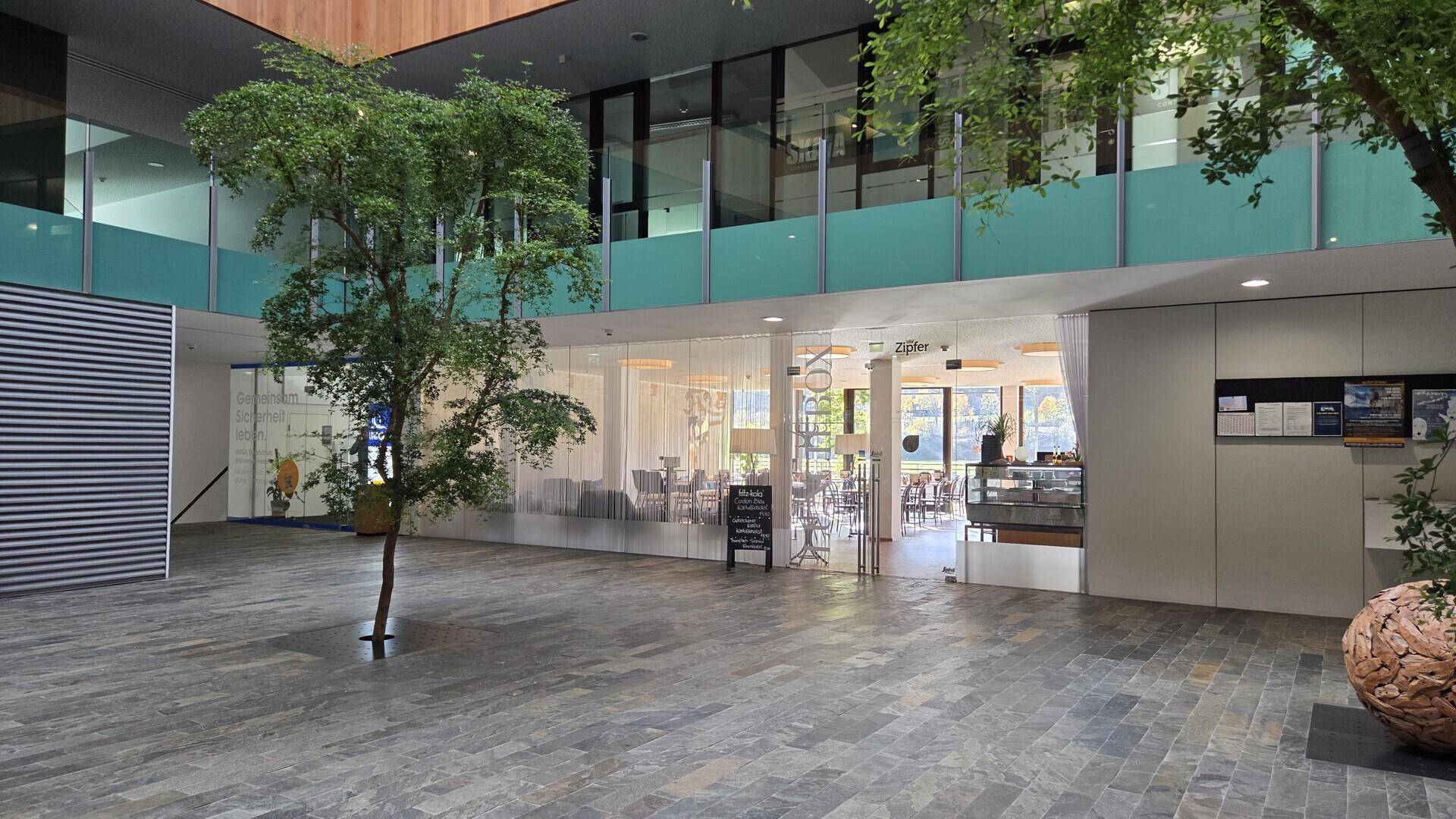 Helle Lobby mit Bäumen, Steinfliesenboden und einem Café mit großen Glasfronten.