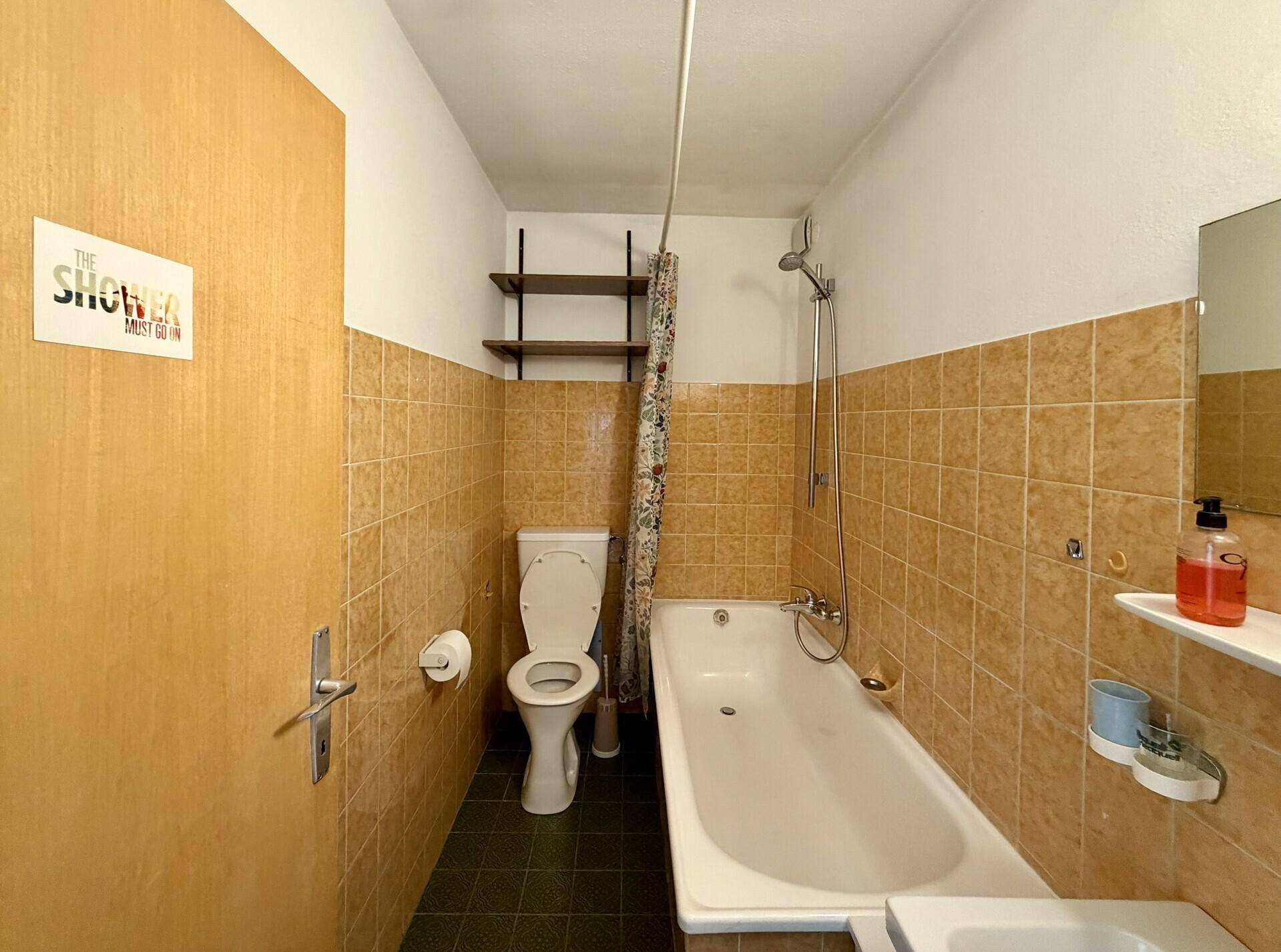 Funktionales Badezimmer mit Badewanne, Duschvorrichtung und braunen Wandfliesen im 80er-Jahre-Stil.