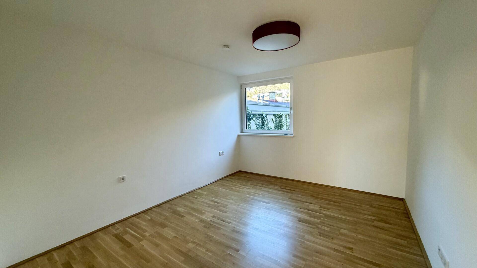 Helles Schlafzimmer mit warmem Parkettboden und einem Fenster, bereit zur individuellen Gestaltung.